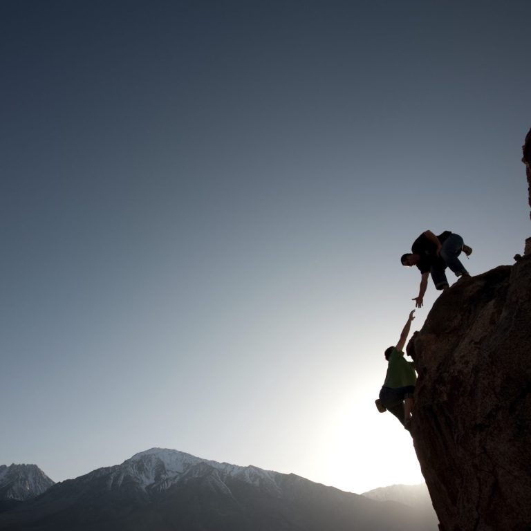 Person on a mountain face, reaching out and about to be pulled up by another person